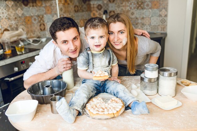 Familieshow van twee ouders en een kind zittend op de keukentafel terwijl ze spelen met bloem en een taart proeven.