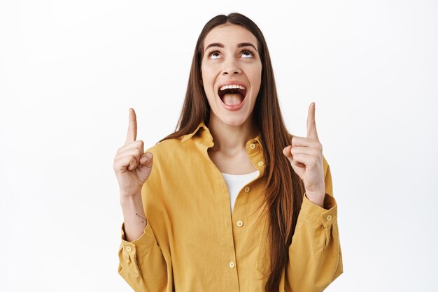 Fantastische aanbieding recht boven Verbaasde lachende vrouw kijkt en wijst omhoog met onder de indruk en opgewonden gezicht en bekijkt iets goeds bovenop in geel shirt tegen witte achtergrond