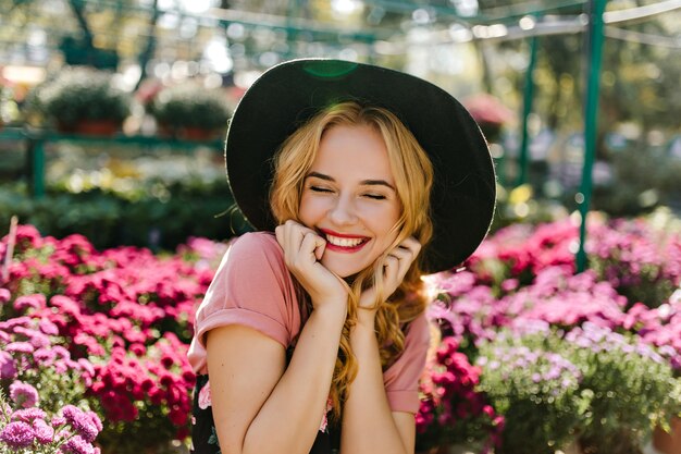 Fascinerend vrouwelijk model met krullend kapsel dat in tuin lacht. Lieve blanke vrouw die oprechte emoties uitdrukt tijdens het poseren met bloemen.