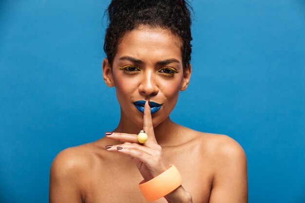 Fascinerende afro-Amerikaanse vrouw die kleurrijke make-up en toebehoren draagt die met vinger op geïsoleerde lippen stellen, over blauwe muur