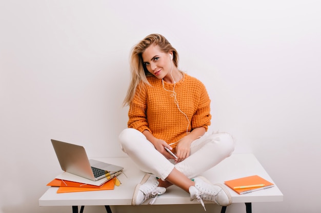 Fascinerende blonde vrouw die in sportschoenen op bureau zit en met speels glimlach na het werk met laptop kijkt