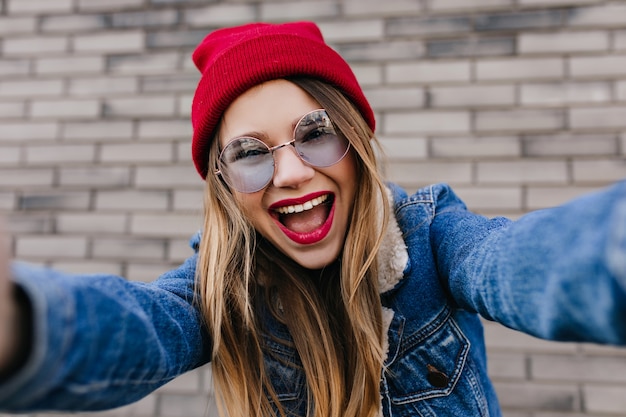 Fascinerende jonge vrouw met lichte make-up selfie maken op bakstenen muur. Foto van dromerig wit vrouwelijk model in blauwe glazen en spijkerjasje dat foto van zich neemt.