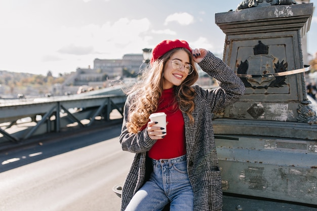 Fascinerende verlegen vrouw in glazen koffie drinken op brug in winderige dag