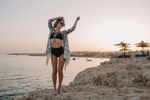 Fasionable model poseren op strand, op zonsondergang, meisje op vakantie. Sexy vrouw, gekleed in zwarte bikini, zwembroek met hoge taille, vest, cape met versieringen.