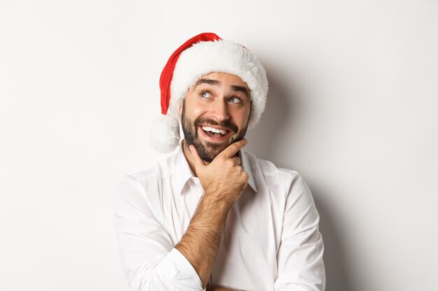 Feest, wintervakantie en feestconcept. Close-up van de gelukkige mens die lijst van de Kerstmisgift, het dragen van santahoed, nadenkend linkerbovenhoek plant