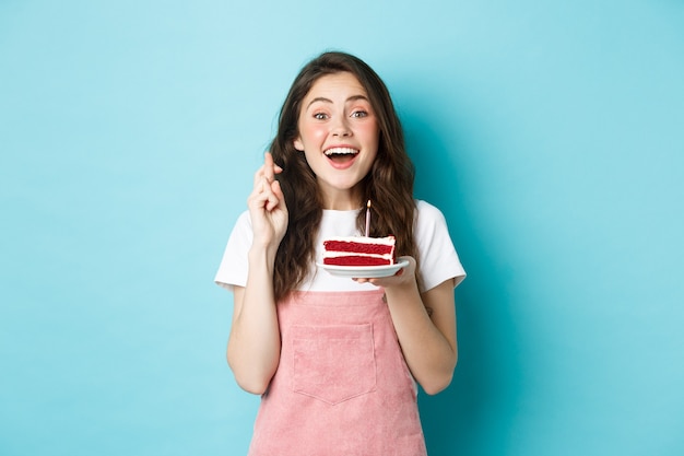 Feestdagen en feest. Opgewonden vrouw die verjaardag viert, vingers kruist en een wens doet terwijl ze een kaars op een taart blaast, staande tegen een blauwe achtergrond