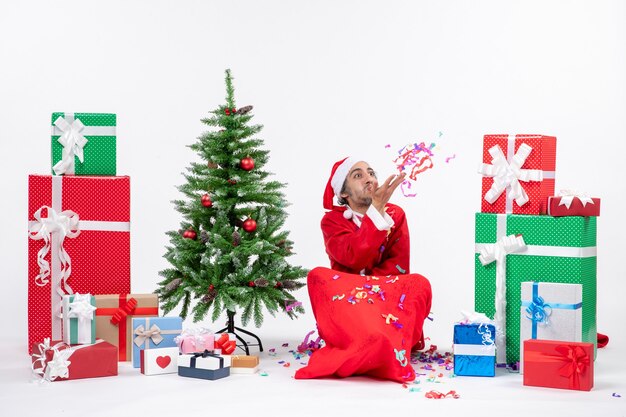 Feestelijke vakantiestemming met grappige kerstman zittend op de grond en spelen met kerstversiering in de buurt van geschenken en versierde kerstboom op witte achtergrond