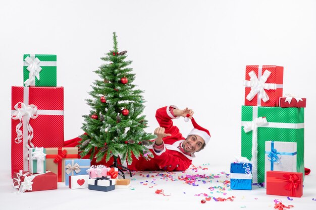 Feestelijke vakantiestemming met jonge positieve kerstman liggend achter kerstboom in de buurt van geschenken op witte achtergrond