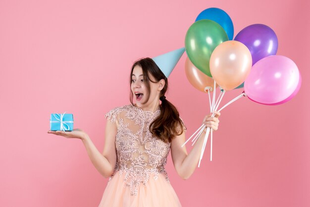 feestmeisje met feestmuts met kleurrijke ballonnen kijken naar cadeau op roze