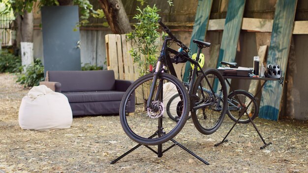 Fiets rustend op een standaard met speciaal gereedschap op maat gemaakt voor nauwkeurige aanpassingen. De omgeving biedt een glimp van de tuin met een kapotte fiets die wacht op reparatie en herstel naar een goede werkende staat.