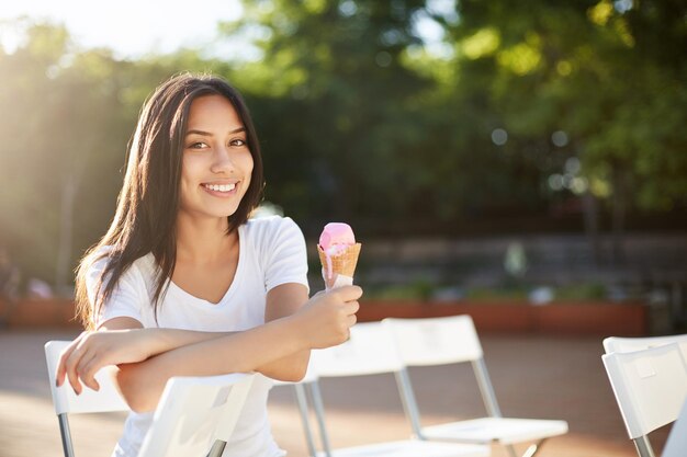 Fijne aziatische vrouw die in het park zit te wachten op een concert dat ijs eet en geniet van haar zomertijd