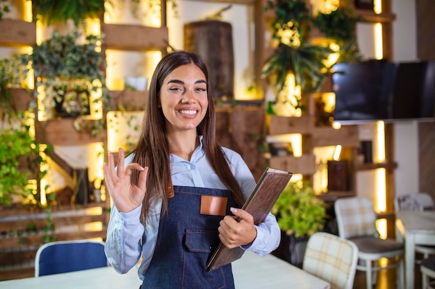 Fijne, mooie glimlachende serveerster met een schort met een mapmenu in een restaurant en kijkend naar een camera die in een gezellig koffiehuis staat, goede service