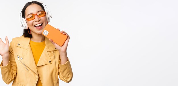 Fijne stijlvolle aziatische vrouw in een zonnebril die muziek luistert in een koptelefoon met smartphone die zingt en danst over een witte achtergrond