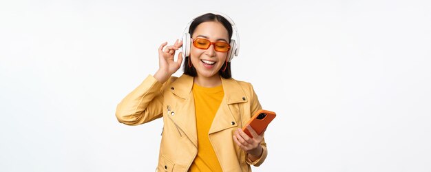 Fijne stijlvolle aziatische vrouw in een zonnebril die muziek luistert in een koptelefoon met smartphone die zingt en danst over een witte achtergrond