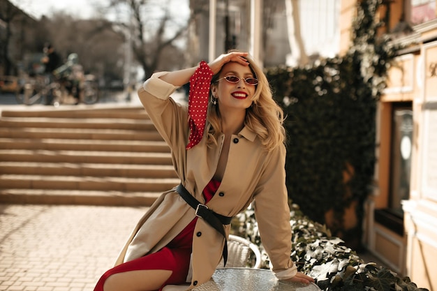 Fijne vrolijke blonde krullende vrouw in zijden heldere jurk, kleurrijke zonnebril en beige trenchcoat zit op tafel en glimlacht naar buiten