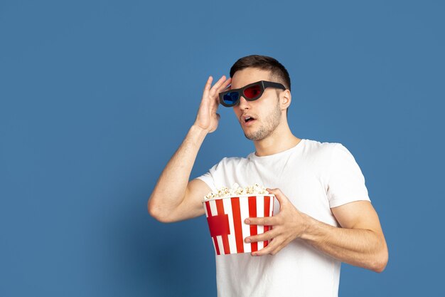 Film kijken met popcorn. Portret van een blanke jongeman op blauwe studiomuur.