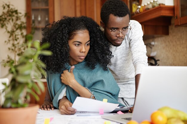 Financiële problemen, gezinsbudget en schulden. Gefrustreerde jonge Afrikaanse man en vrouw laptop pc gebruiken terwijl ze samen papierwerk doen, kosten berekenen, rekeningen in hun moderne keuken beheren