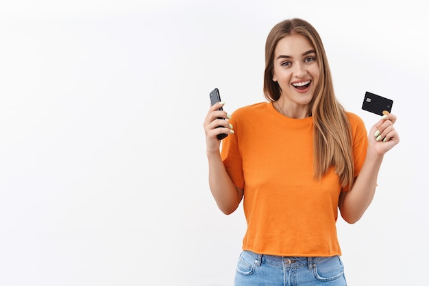 Financiën, geld en banken concept. Portret van een gelukkig, blond meisje kreeg haar salaris, kocht nieuwe kleren online winkelend, hield mobiele telefoon en creditcard vast en glimlachte breed