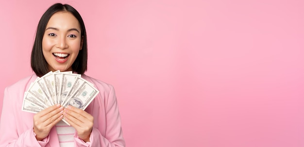 Financiën microkrediet en mensenconcept gelukkige glimlachende aziatische onderneemster die dollarsgeld toont die zich in kostuum tegen roze achtergrond bevinden Premium Foto