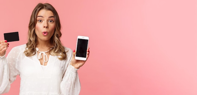 Financiën winkelen en technologie concept close-up portret van opgewonden blond schattig meisje in witte jurk vouwen lippen geamuseerd blik camera als weergegeven: creditcard en mobiele telefoon roze achtergrond