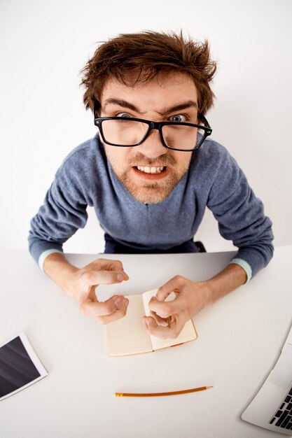 Fish-eye shot van verontruste, geïrriteerde en pissige man die zijn handen balde, boos staarde met gebalde tanden, fronsend fronsend, aan bureau zit