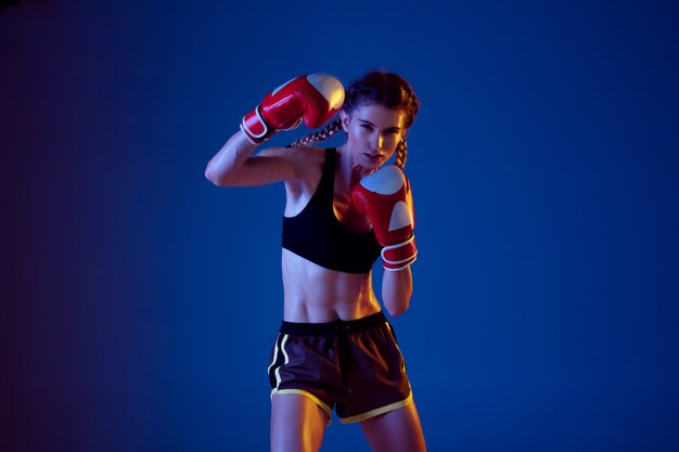 Fit blanke vrouw in sportkleding boksen op blauwe achtergrond in neonlicht.