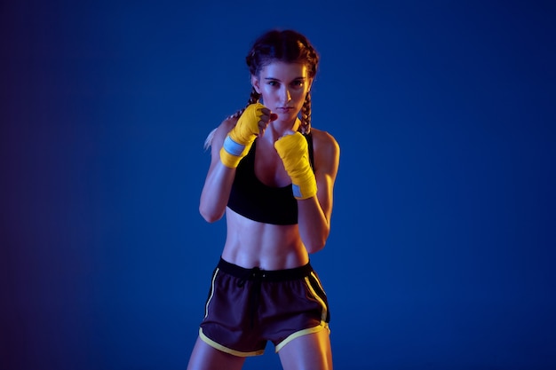 Fit blanke vrouw in sportkleding boksen op blauwe achtergrond in neonlicht.