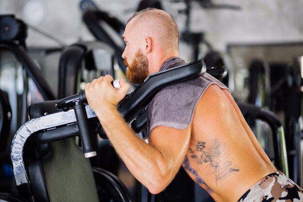 Fit getatoeëerde bebaarde man doet squats in een trainingsmachine