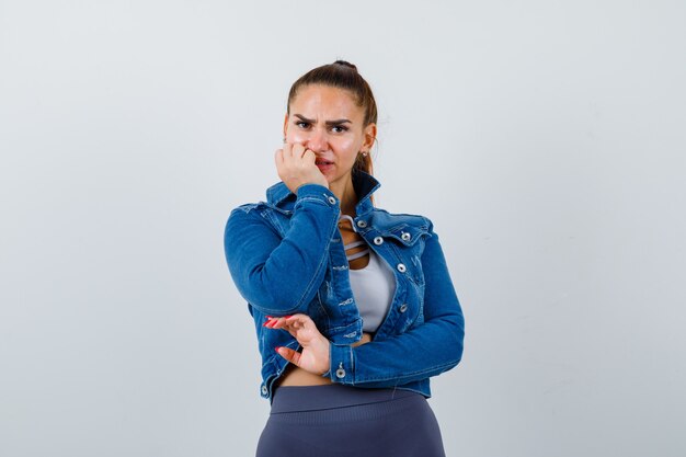 Fit vrouw bijten vingers emotioneel in crop top, spijkerjasje, legging en angstig kijken, vooraanzicht.