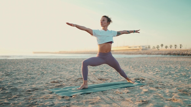 Fit vrouw doet yoga op mat aan zee Mooi yogi meisje in sportkleding staande in krijger yoga pose op het strand