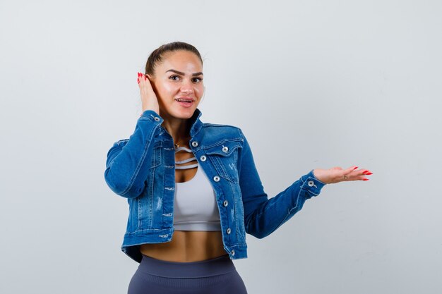 Fit vrouw in crop top, spijkerjasje, legging die hand op oor drukt, palm opzij spreidt en er gelukkig uitziet, vooraanzicht.