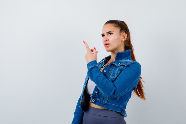 Fit vrouw in crop top, spijkerjasje, legging die met de wijsvinger omhoog wijst en er gefocust uitziet, vooraanzicht.