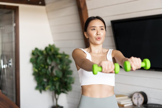 Fitness Aziatisch meisje doet thuis training in de woonkamer en tilt halters op met activewear voor spo...