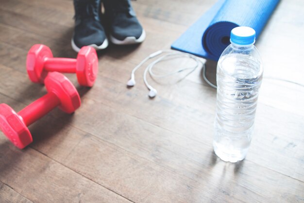 Fitness concept met fles water, sneakers, rode halters, yoga mat en koptelefoon op houten vloer, kopieer ruimte