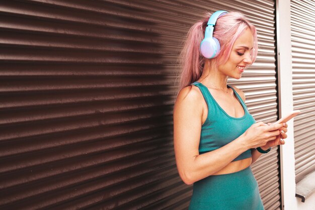 Fitness lachende vrouw in groene sportkleding met roze haar Jonge mooie model met perfect lichaam Vrouw in de straat in de buurt van rolluikmuur Muziek luisteren in draadloze hoofdtelefoons