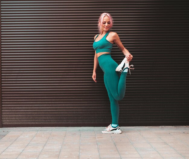 Fitness lachende vrouw in groene sportkleding met roze haar Jonge mooie model met perfect lichaam Vrouwelijke poseren in de straat in de buurt van rolluik muur die zich uitstrekt voor de opleiding