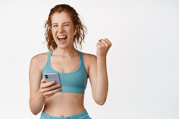 Fitness meisje bereikt sport app doel op mobiele telefoon schreeuwen tevreden en glimlachend blij training in activewear staande tegen een witte achtergrond