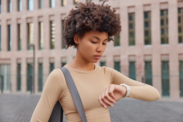 Fitness vrouw controleert resultaten op smartwatch gekleed in beige sportkleding gedragen opgerolde karemat gaat regelmatig sporten poseert in de buurt van stedelijk gebouw buitenshuis Atletische vrouw na het sporten