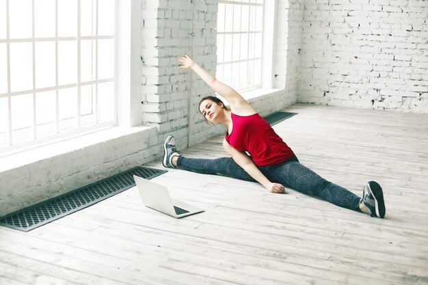 Flexibiliteit en kracht. Prachtige jonge vrouw met perfect lichaam spieren thuis uitrekken, zijbeen splitsen en naar rechts buigen tijdens het kijken naar yoga tutorial online op draagbare computer