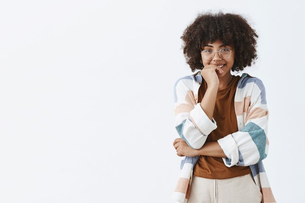 flirterig schattig donkerhuidig vrouwelijk model met krullend kapsel in transparante glazen en trendy outfit breed glimlachend met verlegen uitdrukking die mond bedekt met palm