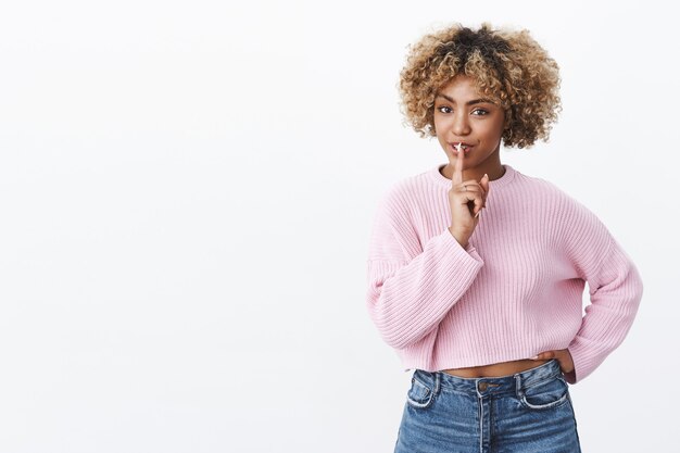 Flirterige Afro-Amerikaanse vrouw met blond kapsel die er sensueel uitziet met een shh-gebaar met een lichte romantische grijns, hand op taille en wijsvinger over mond, geheim verbergend of verrassing makend