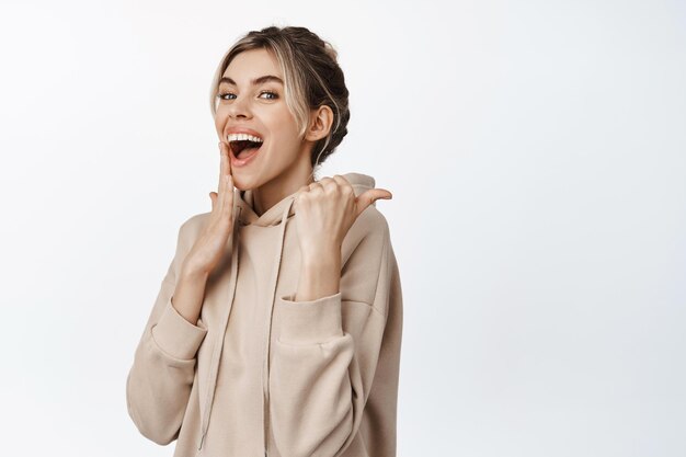 Flirterige jonge vrouw die met de vinger naar rechts wijst en koket lacht in beige hoodie tegen een witte achtergrond Advertentieconcept