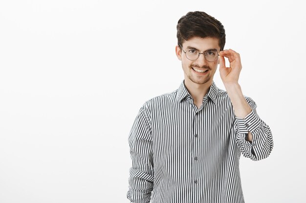 flirterige nerdy kerel in ronde bril met baard en snor, die de rand van een bril vasthoudt en breed lacht, zich zelfverzekerd en ontspannen voelt na het zeggen van ophaallijn tegen aantrekkelijk meisje in de bar