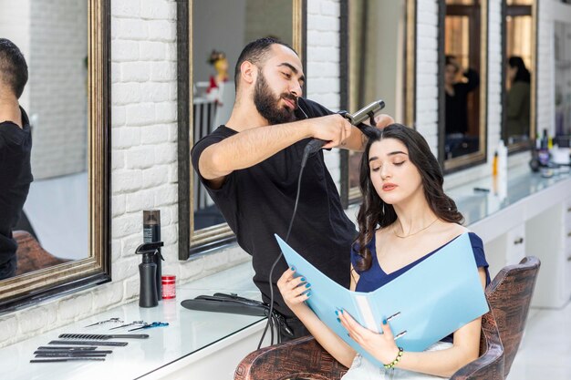 Foto bij de kapperszaak Jonge kapper probeert het haar van haar klanten te vormen en de klant kijkt naar de catalogus Foto van hoge kwaliteit