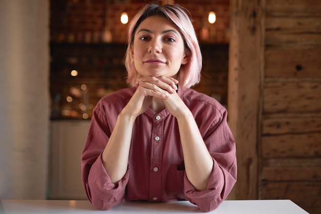 Foto of stijlvolle zelfverzekerde jonge 20-jarige vrouw copywriter draagt katoenen shirt en neusring met gevouwen handen onder de kin, lachend, werkend vanuit huis, comfortabel zittend aan een witte tafel