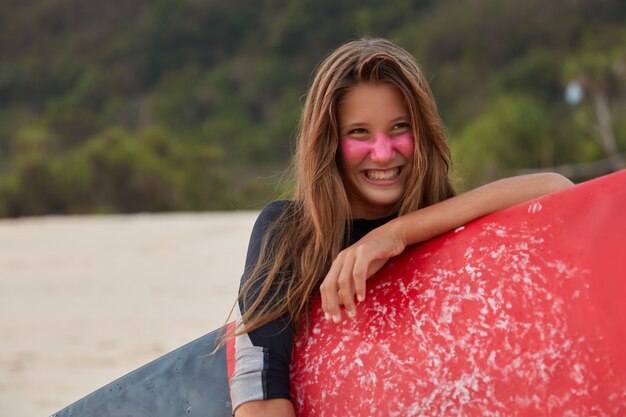 Foto van aangenaam ogende Europese vrouw met gelukkige uitdrukking, verheugt zich over de dag om te surfen, houdt gewaxte surfplank vast, dol op watersport, draagt duikpak, wil onderste bocht laten zien. Horizontaal schot.