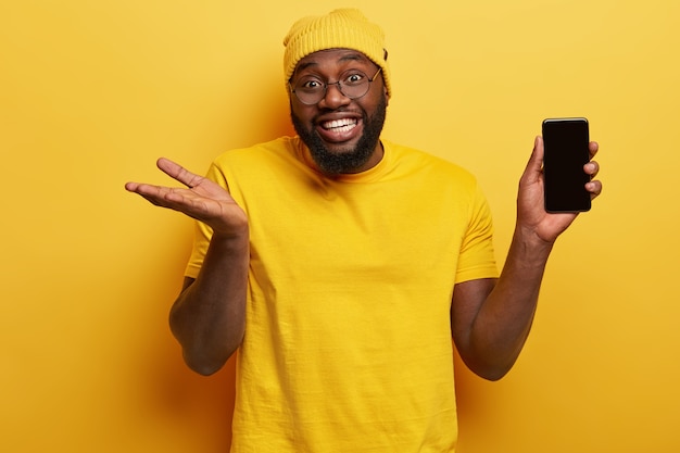 Foto van aantrekkelijke donkere man met brede glimlach, witte tanden, houdt slimme telefoon met leeg scherm voor uw informatie, draagt een ronde bril, werpt handpalm met onzekerheid, neemt een beslissing