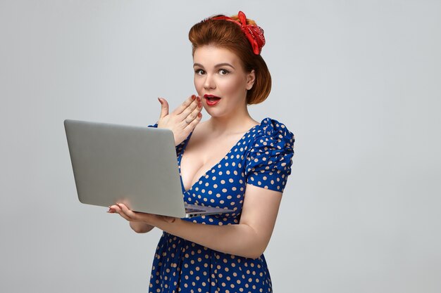 Foto van aantrekkelijke glamoureuze jonge vrouw, gekleed in pin-up stijl jurk en retro kapsel met geschokte verbaasde uitdrukking, mond bedekt, generieke laptopcomputer open voor haar