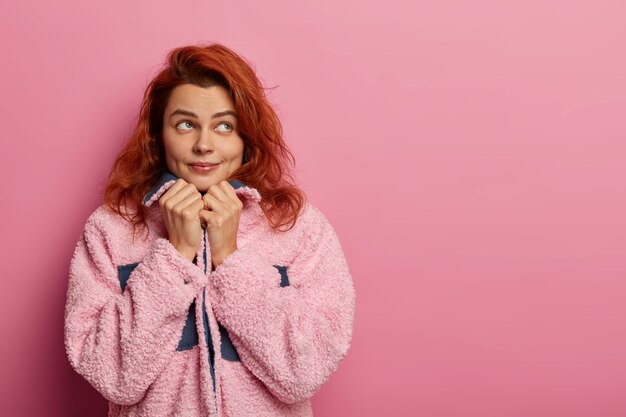 Foto van aantrekkelijke roodharige vrouw houdt handen onder de kin en kijkt bedachtzaam opzij, stelt zich voor tijdens vrije tijd buiten lopen, draagt winterjas