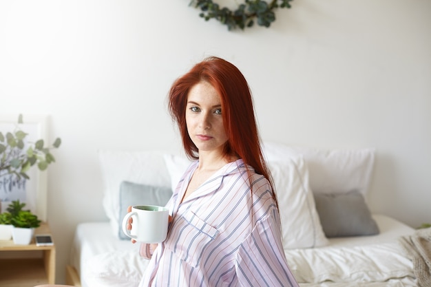 Foto van aantrekkelijke slaperige jonge roodharige Europese vrouw stijlvolle gestreepte nacht pak dragen, zittend in haar gezellige slaapkamer, verse sterke koffie drinken vroeg in de ochtend voor het werk